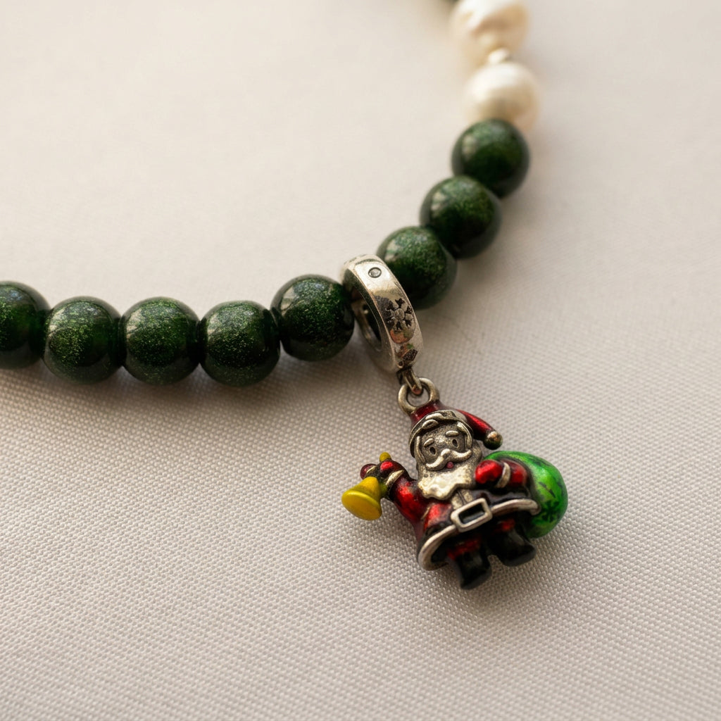 Close-up of a sterling silver Santa charm with a yellow bell and green sack beside sparkling green sandstone beads on cream fabric.