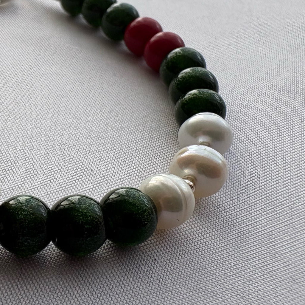 Close-up of three lustrous white pearls between sparkling dark green sandstone beads, with red coral accents on pale fabric.