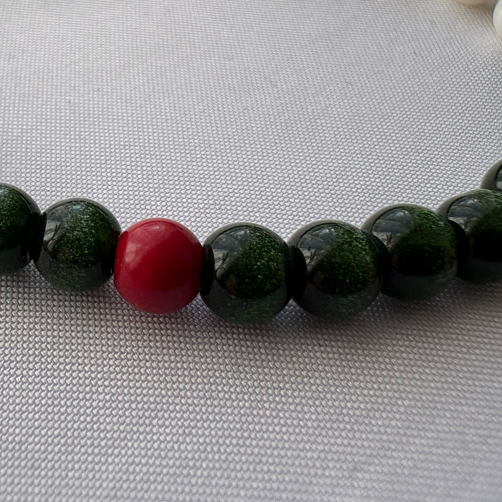Close-up of a glossy red coral bead among sparkling dark green sandstone beads on pale woven fabric.