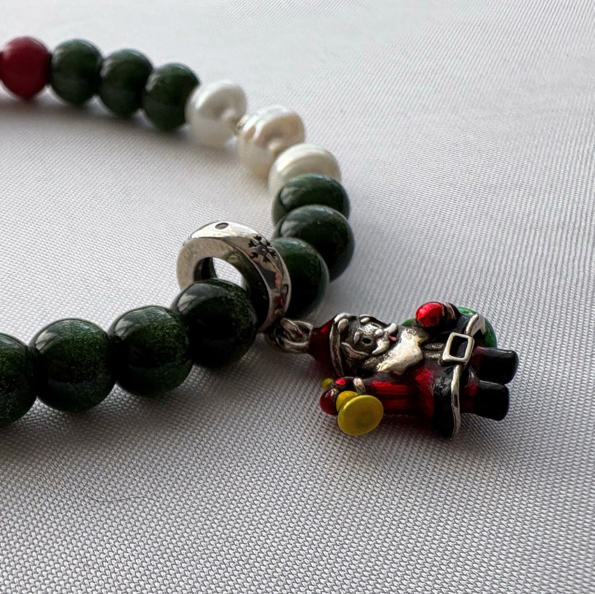 Close-up of an enamel Santa charm holding a yellow bell beside green sandstone beads and three white pearls on pale fabric.