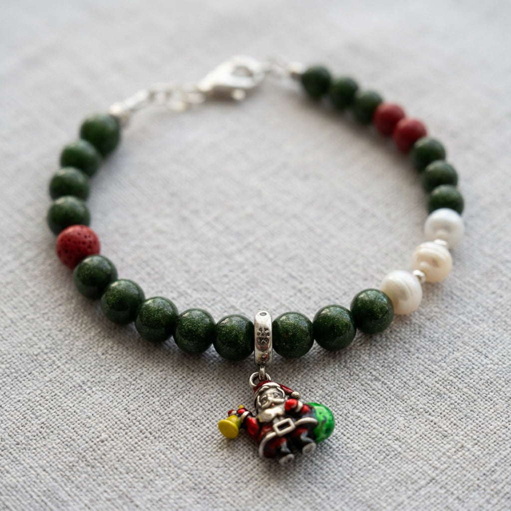 Green sandstone bracelet with red coral accents, three white pearls and an enamel Santa charm, arranged on grey linen.
