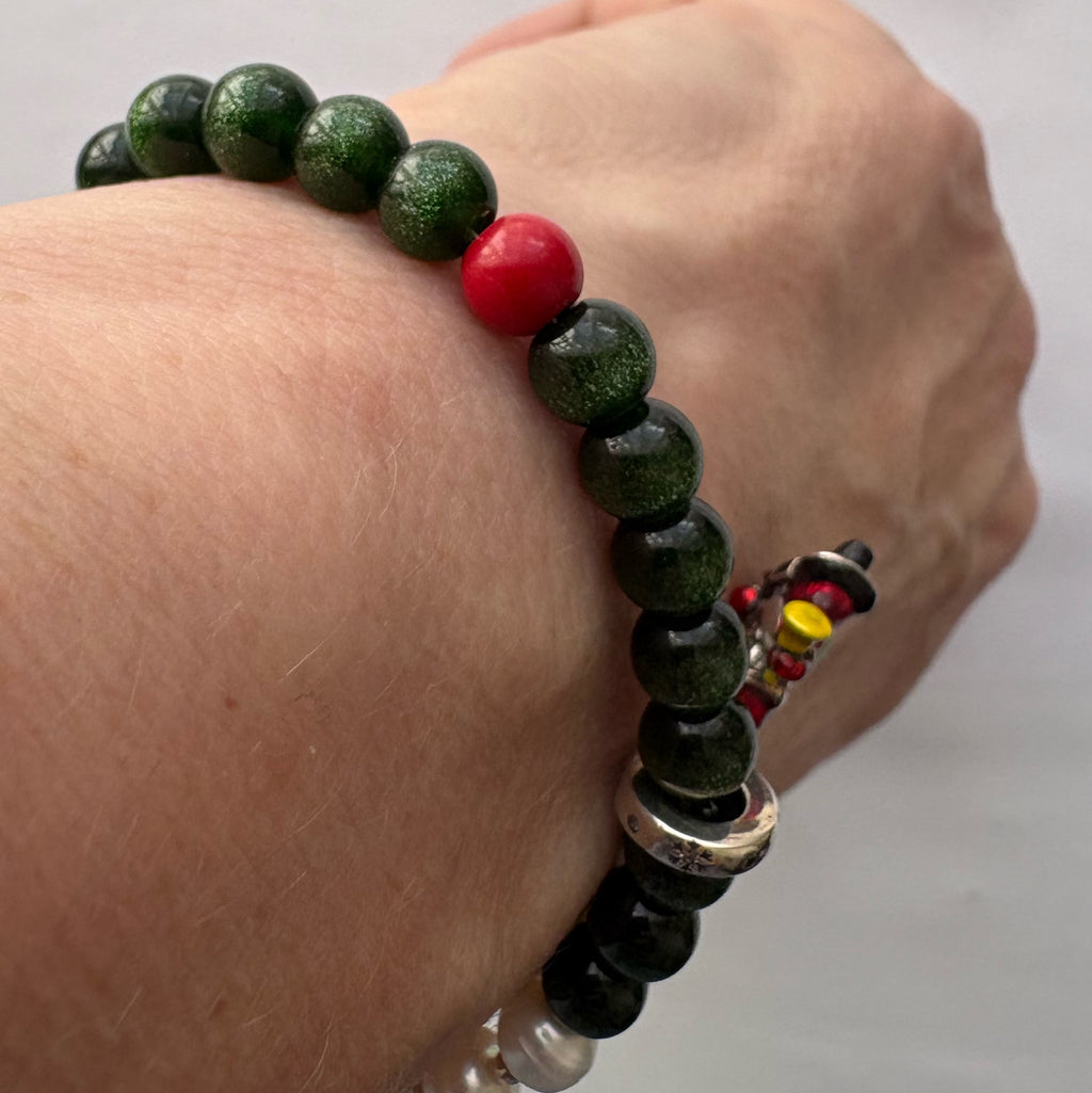 Angled wrist view of a sparkling green sandstone bracelet with a red coral accent and enamel Santa charm to one side.