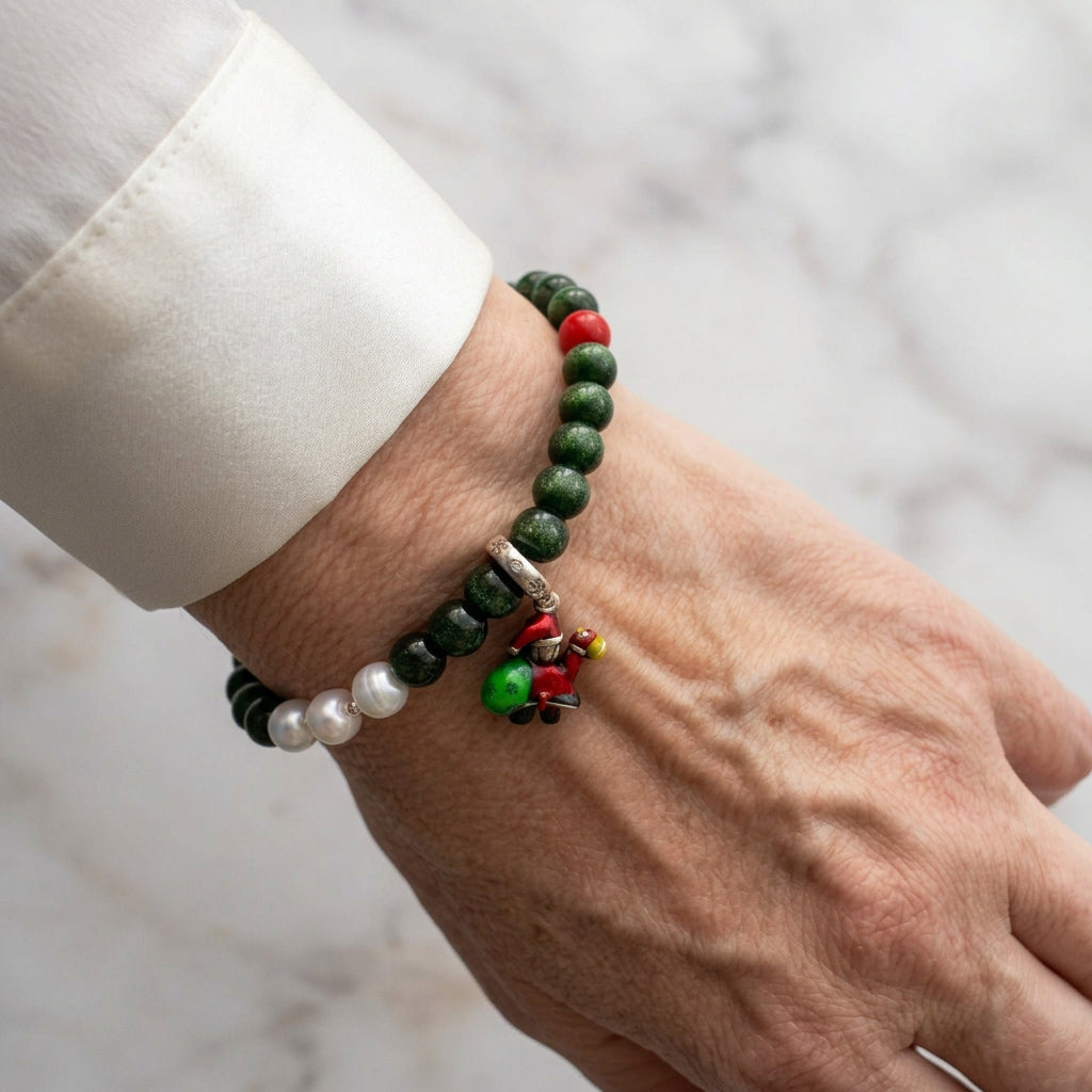 Deep green sandstone bracelet with red coral and three white pearl accents, featuring a colourful Santa charm, worn on the wrist beside a white silk shirt cuff.