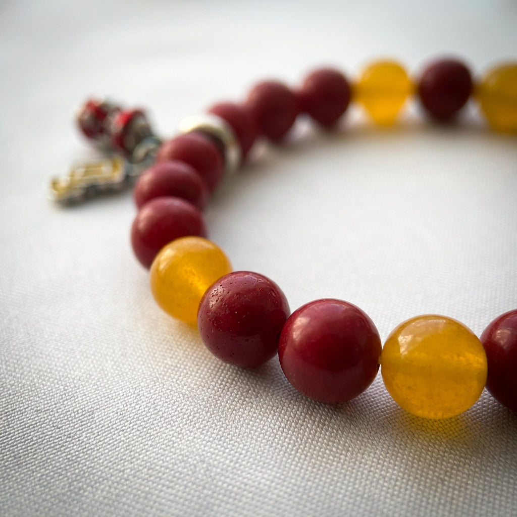 Close-up of glossy red coral and translucent yellow topaz beads on white fabric, with Christmas charms softly blurred behind.⠋