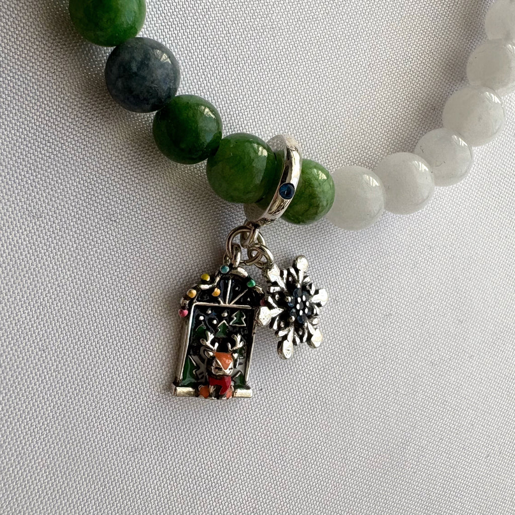 Close-up of reindeer Advent door and snowflake charms hanging from a white and green jade bracelet on pale fabric.