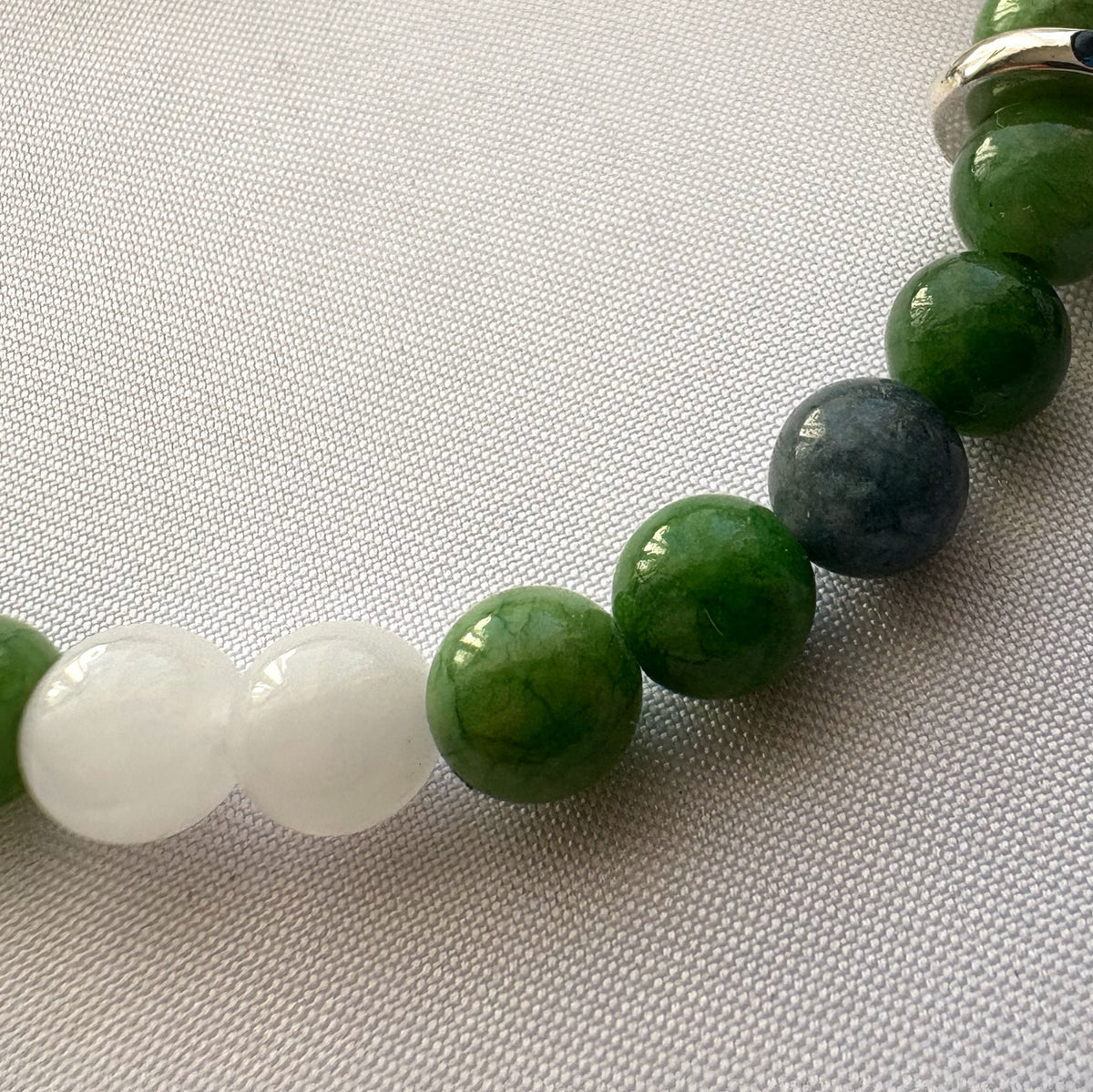 Close-up of polished white, green and dark grey beads on a jade bracelet resting on pale woven fabric.