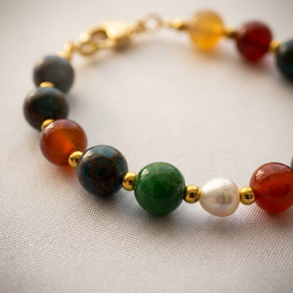 Close-up of blue jasper, green jade, a white baroque pearl and orange-red carnelian beads separated by gold-coloured spacers on pale fabric.