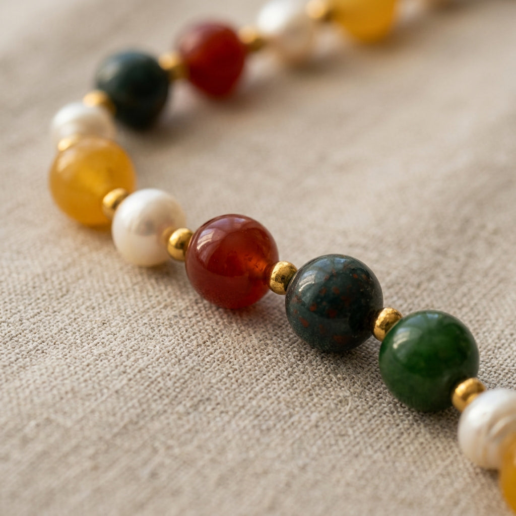 Beaded bracelet on cream linen, with a green jade bead between two white baroque pearls at the front, surrounded by blue jasper, orange-red carnelian, yellow topaz and gold-coloured spacers.