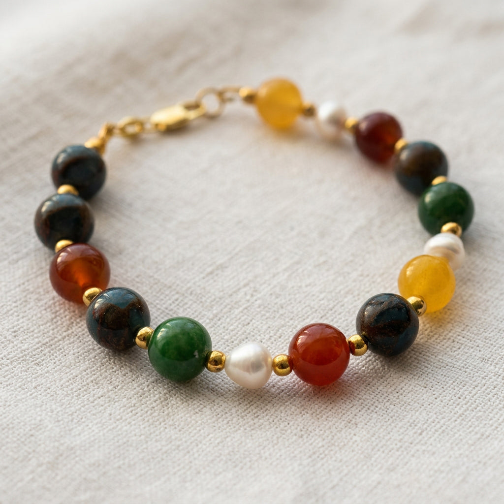 Close-up of orange-red carnelian, blue jasper, green jade, yellow topaz and white baroque pearls separated by gold-coloured spacers on cream linen.