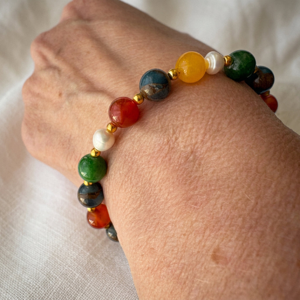 Beaded bracelet worn across the top of a wrist, showing orange-red carnelian, blue jasper, yellow topaz, green jade and white baroque pearls with gold-coloured spacers.