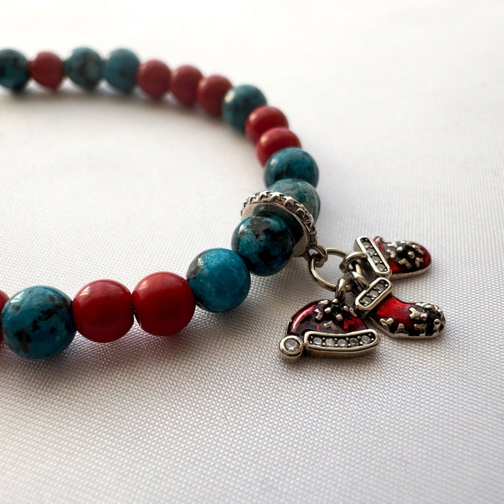 Close-up of red enamel Santa hat and stocking charms beside blue agate and red coral beads on white fabric.