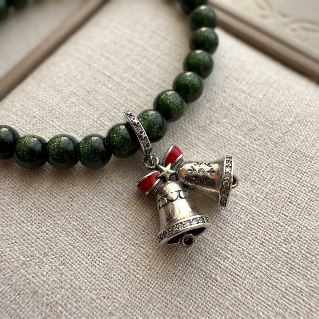 Close-up of twin silver-coloured bell charms with a red bow on a sparkling green sandstone bracelet, resting on beige linen.