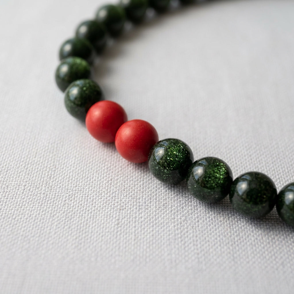 Close-up of two red coral beads among sparkling dark green sandstone beads on white fabric.