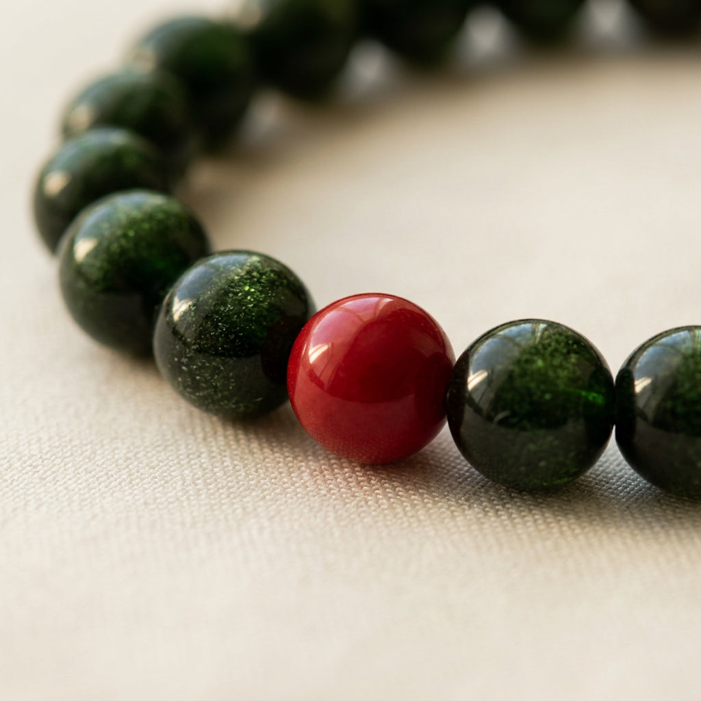 Macro view of a glossy red coral bead between sparkling dark green sandstone beads on cream fabric.