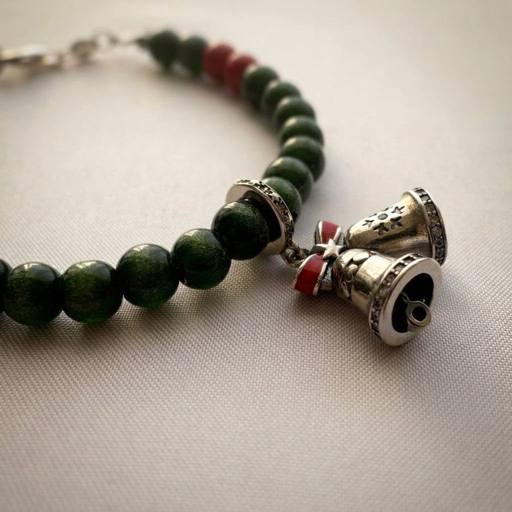 Angled close-up of twin bell charms with a red bow on a green sandstone and red coral bracelet, resting on cream fabric.
