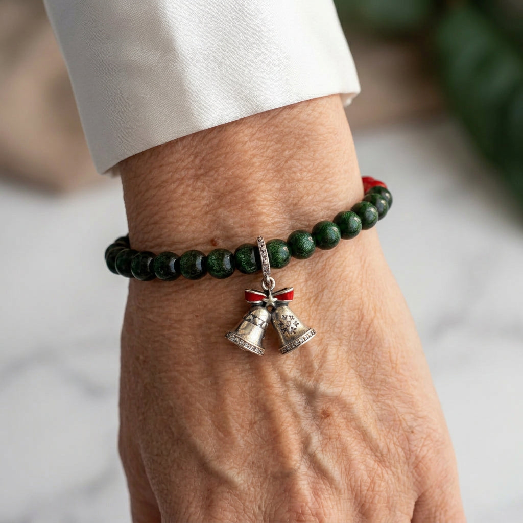 Deep green sandstone bracelet with red coral accents and a silver-coloured double bell charm topped with a red enamel bow, worn on the wrist beside a white silk shirt cuff.