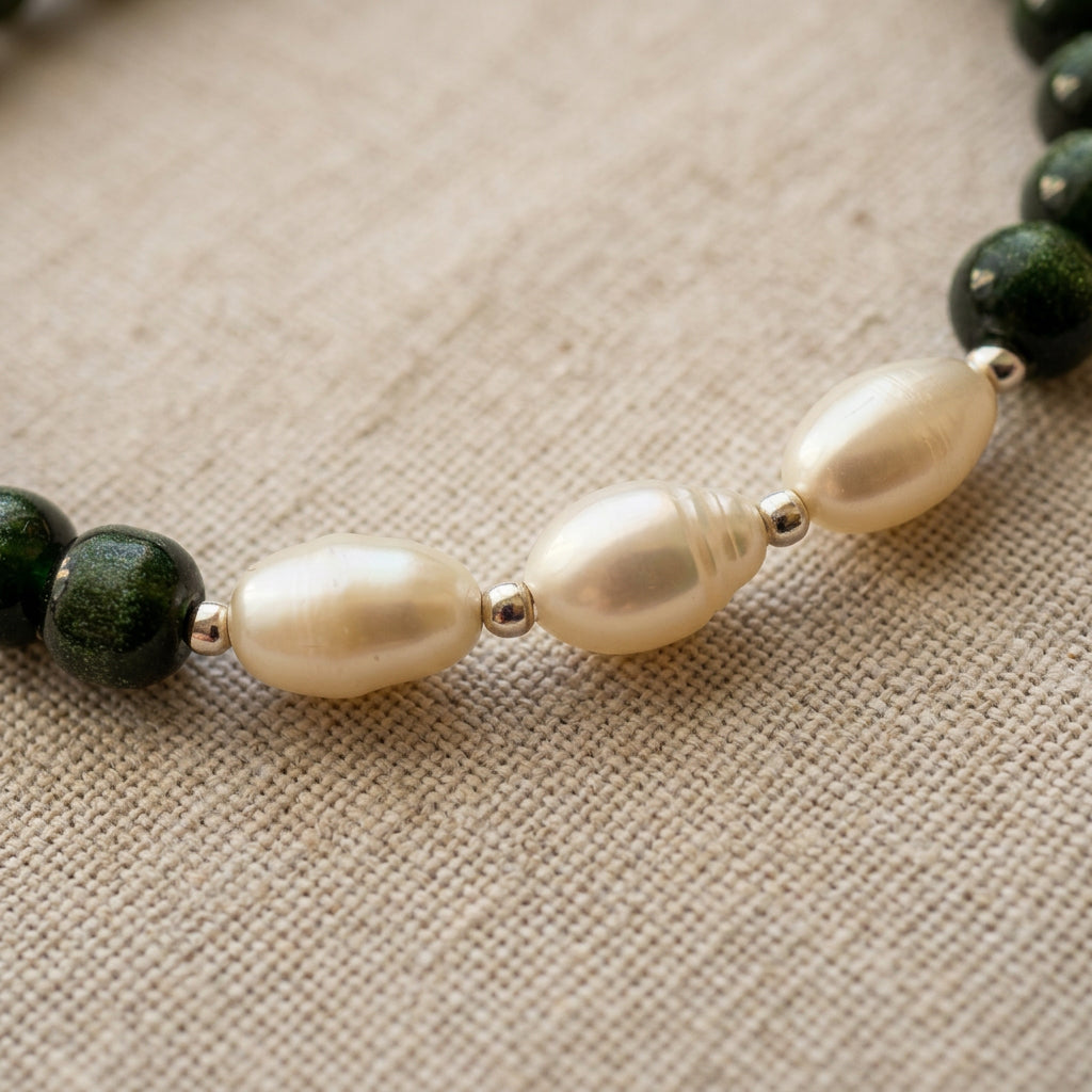 Close-up of three oval white pearls with silver-coloured spacers between dark green sandstone beads on beige linen.