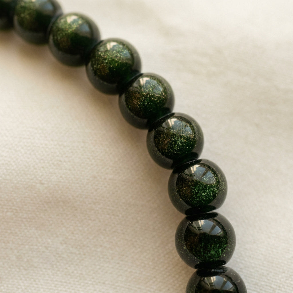 Close-up of glossy dark green sandstone beads with shimmering flecks, separated by black spacers on cream fabric.