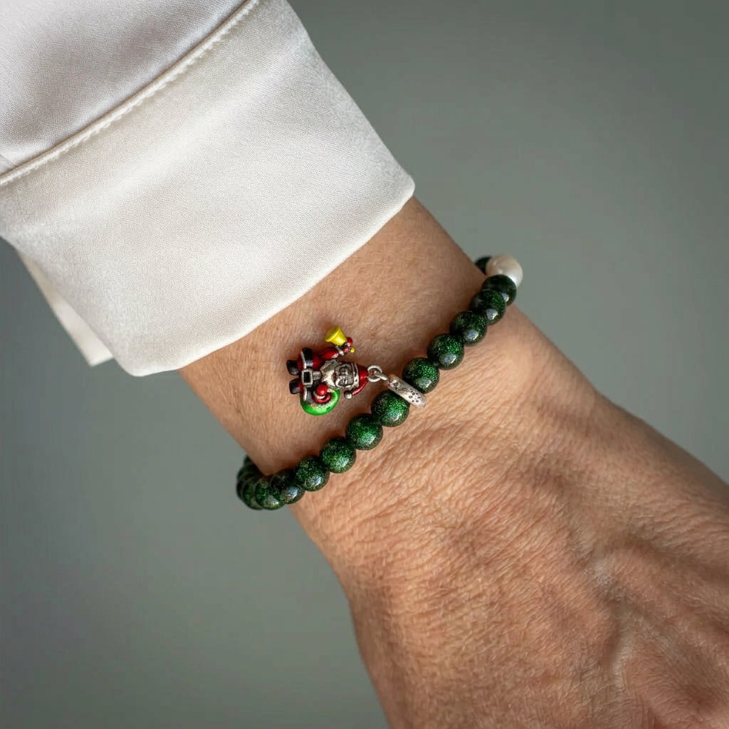 Deep green sandstone bracelet with white pearl accents and an enamel Santa charm holding a yellow bell and green gift sack, worn beside a white silk shirt cuff.