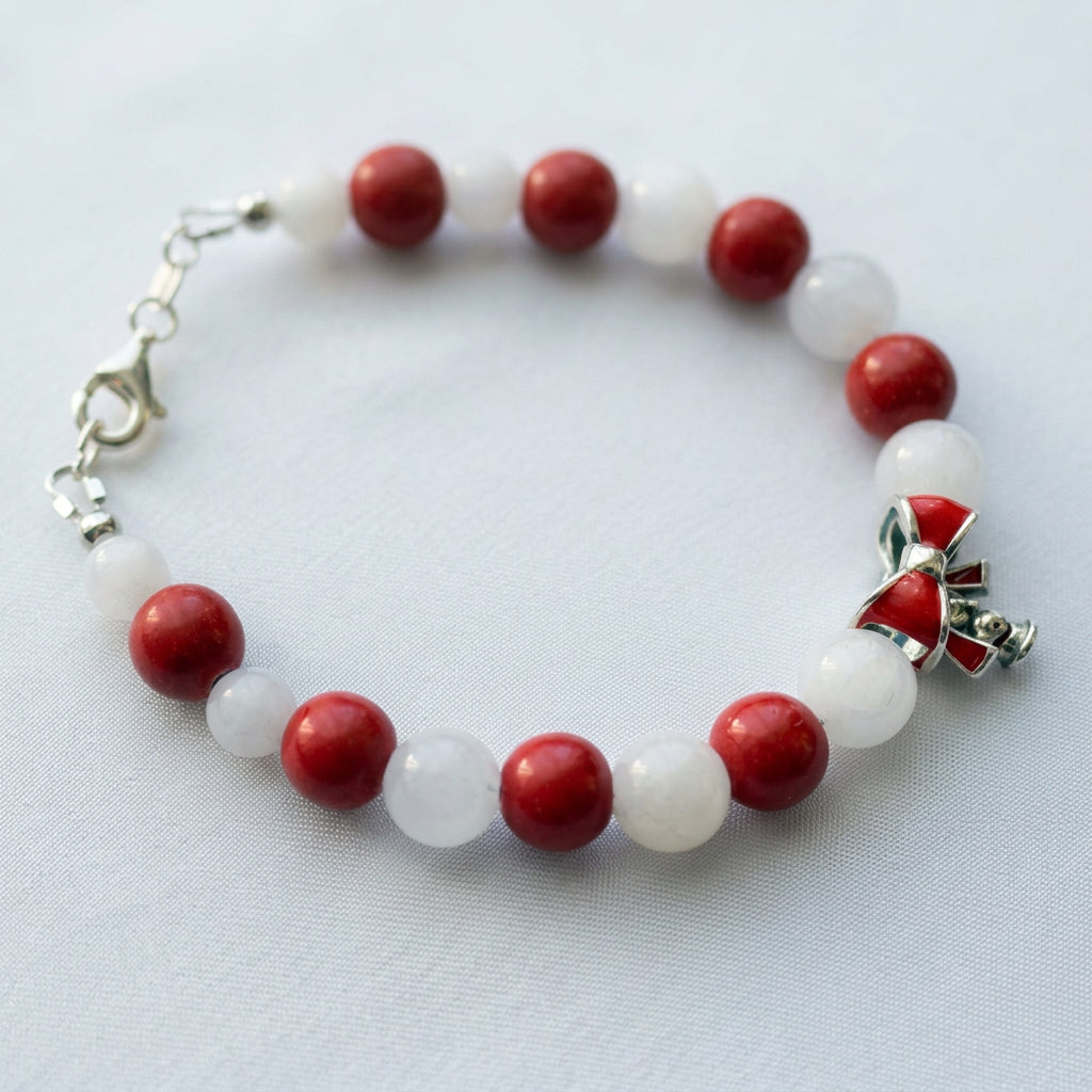 Red coral and white jade bracelet with alternating round beads and a red enamel bow charm, arranged on white fabric.