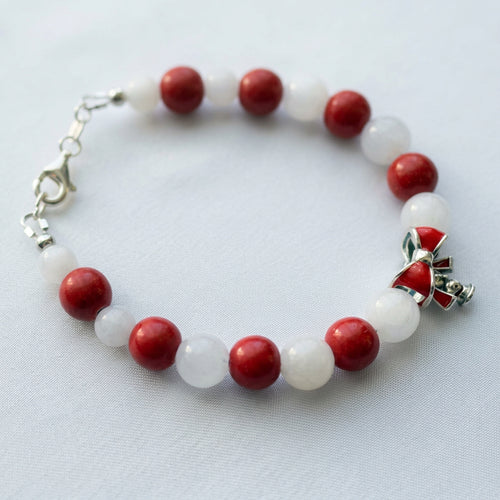Red coral and white jade bracelet with alternating round beads and a red enamel bow charm, arranged on white fabric.