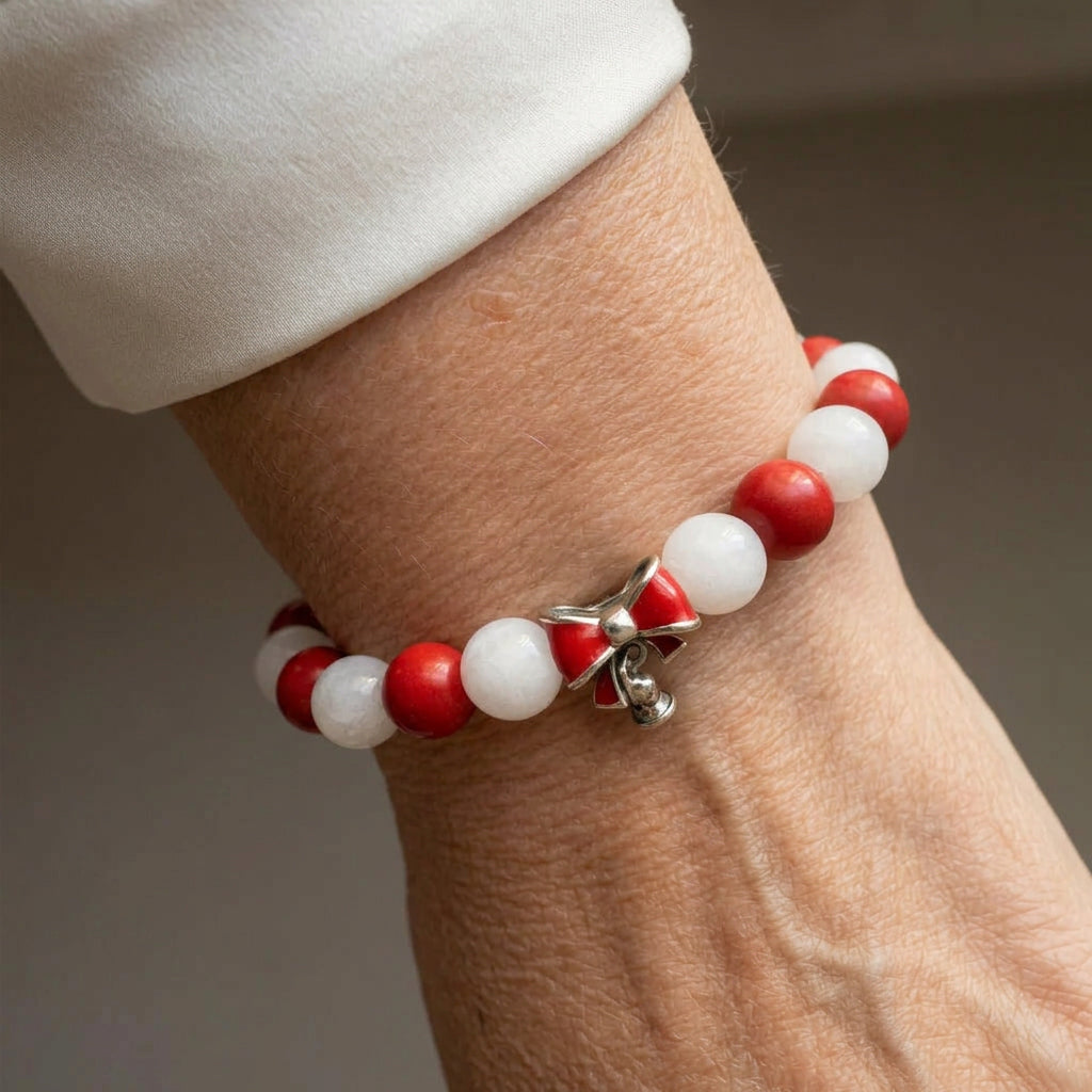 Alternating red coral and white jade bead bracelet with a red enamel bow and small silver-coloured bell charm, worn on the wrist beside a white silk shirt cuff.