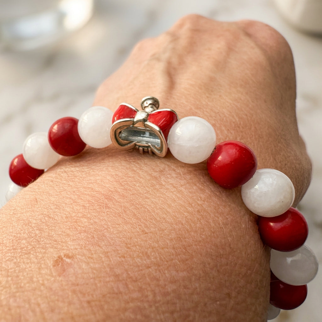 Red coral and white jade bracelet worn on a wrist, showing the back of its red enamel bow charm and a small dangling bell.