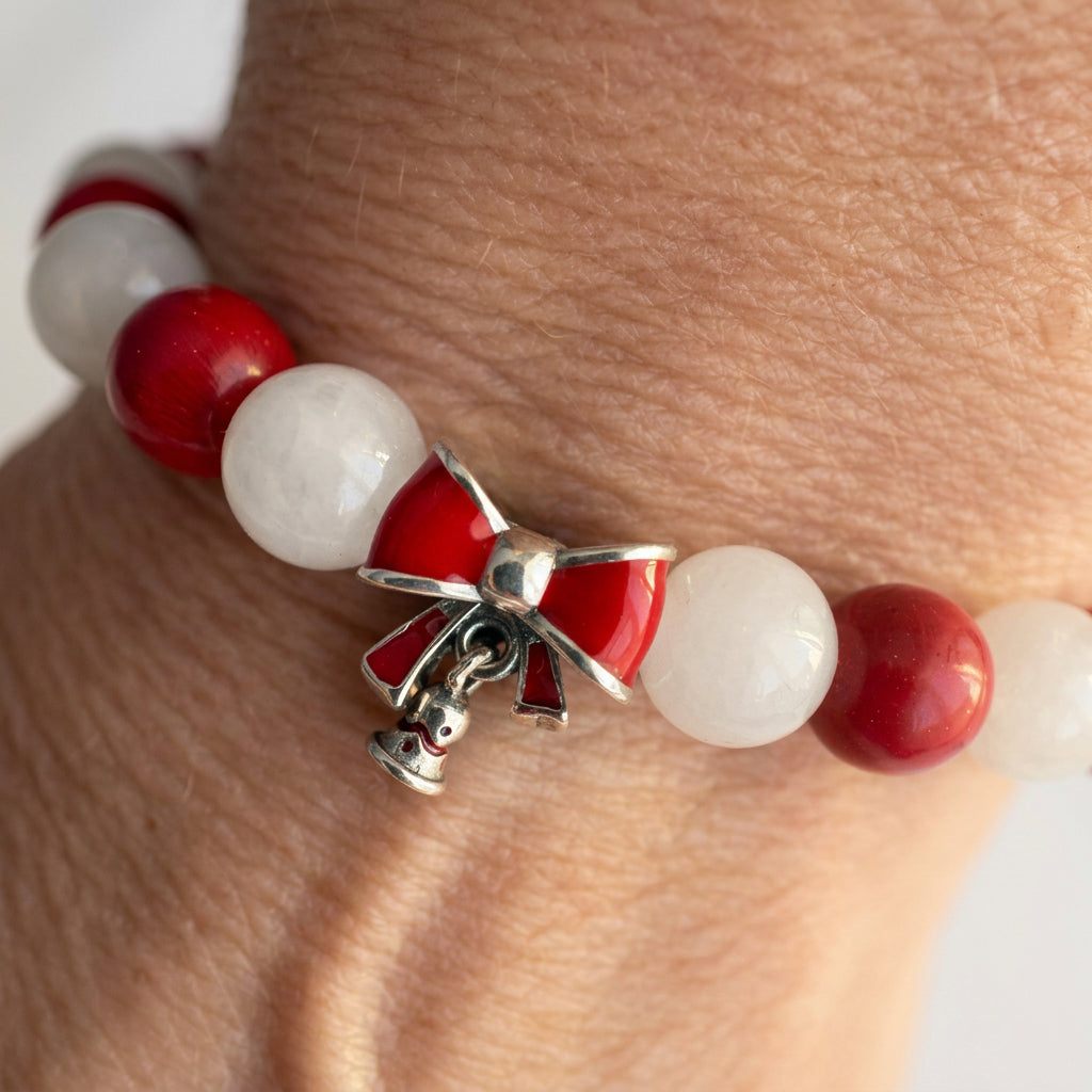 Close-up of a red enamel bow charm with a dangling bell on a red coral and white jade bracelet worn on a wrist.⠋