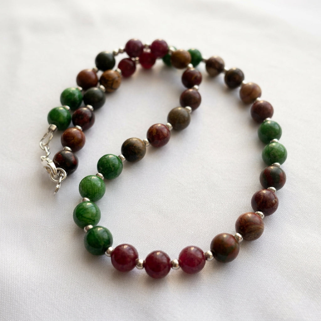 Close-up of a coiled necklace on pale fabric, showing green jade, earthy brown-red jasper and burgundy garnet beads separated by silver-coloured spacers.