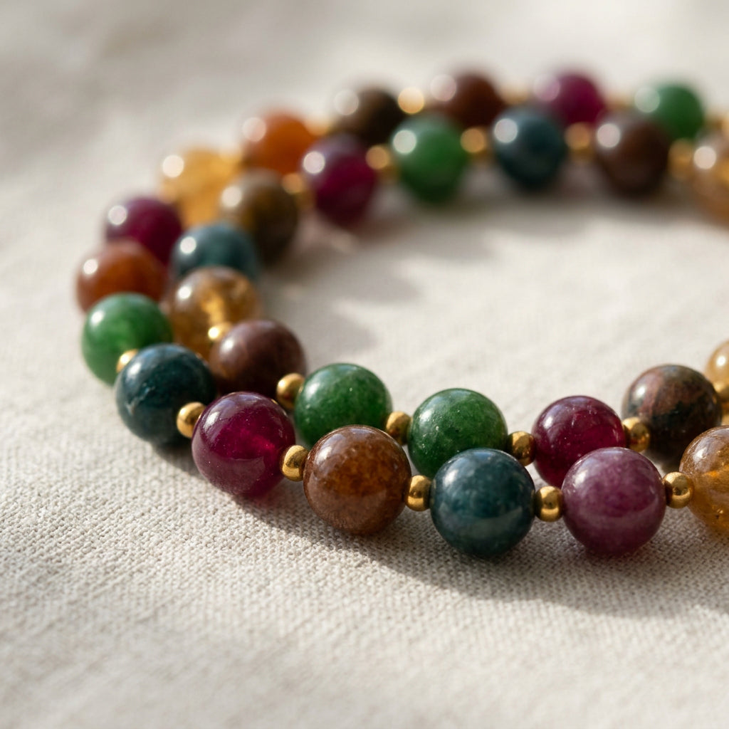 Beaded necklace coiled in overlapping loops on cream linen, showing green, burgundy, blue-grey, brown and amber stones separated by gold-coloured spacers.