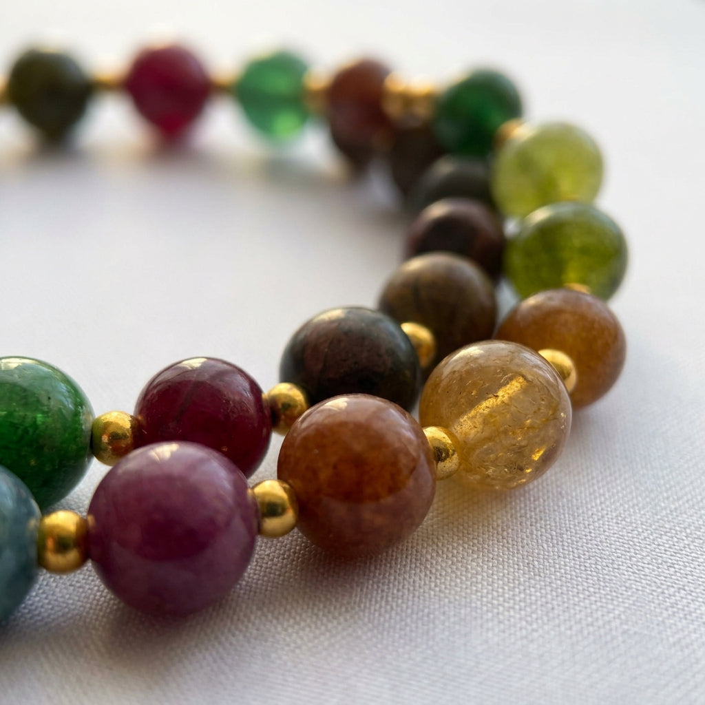 Close-up of a coiled beaded necklace on pale fabric, showing burgundy, earthy brown, translucent amber and green stones separated by small gold-coloured spacers.
