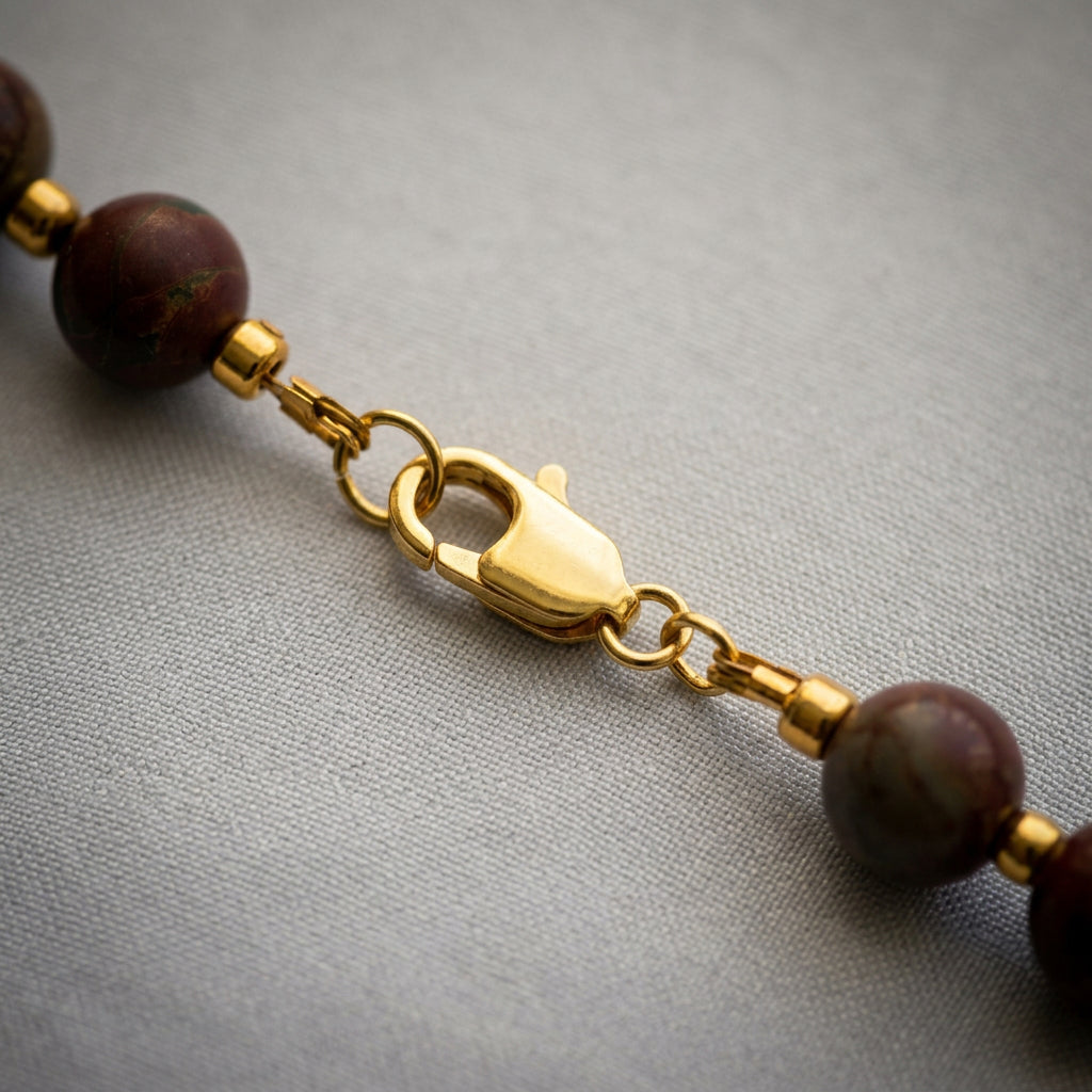 Close-up of the gold-filled lobster clasp and connecting rings between earthy red-brown jasper beads, resting on pale linen.
