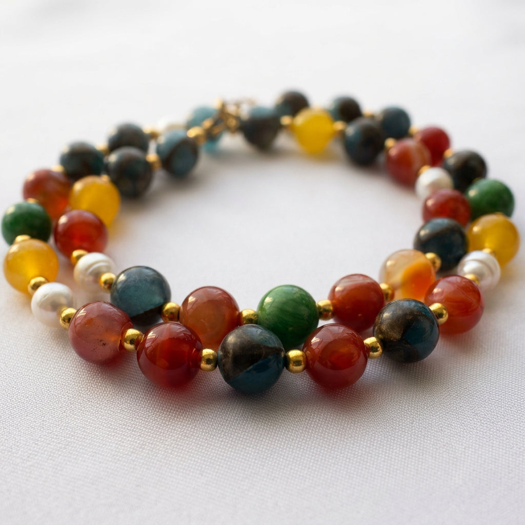 Colourful beaded necklace loosely coiled on pale fabric, showing blue, orange-red, yellow and green stones, white baroque pearls and gold-coloured spacers.