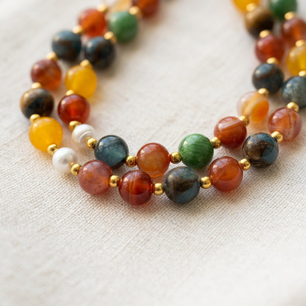 Close-up of a colourful necklace coiled on cream linen, showing translucent orange-red, blue, green and yellow beads, white baroque pearls and gold-coloured spacers.