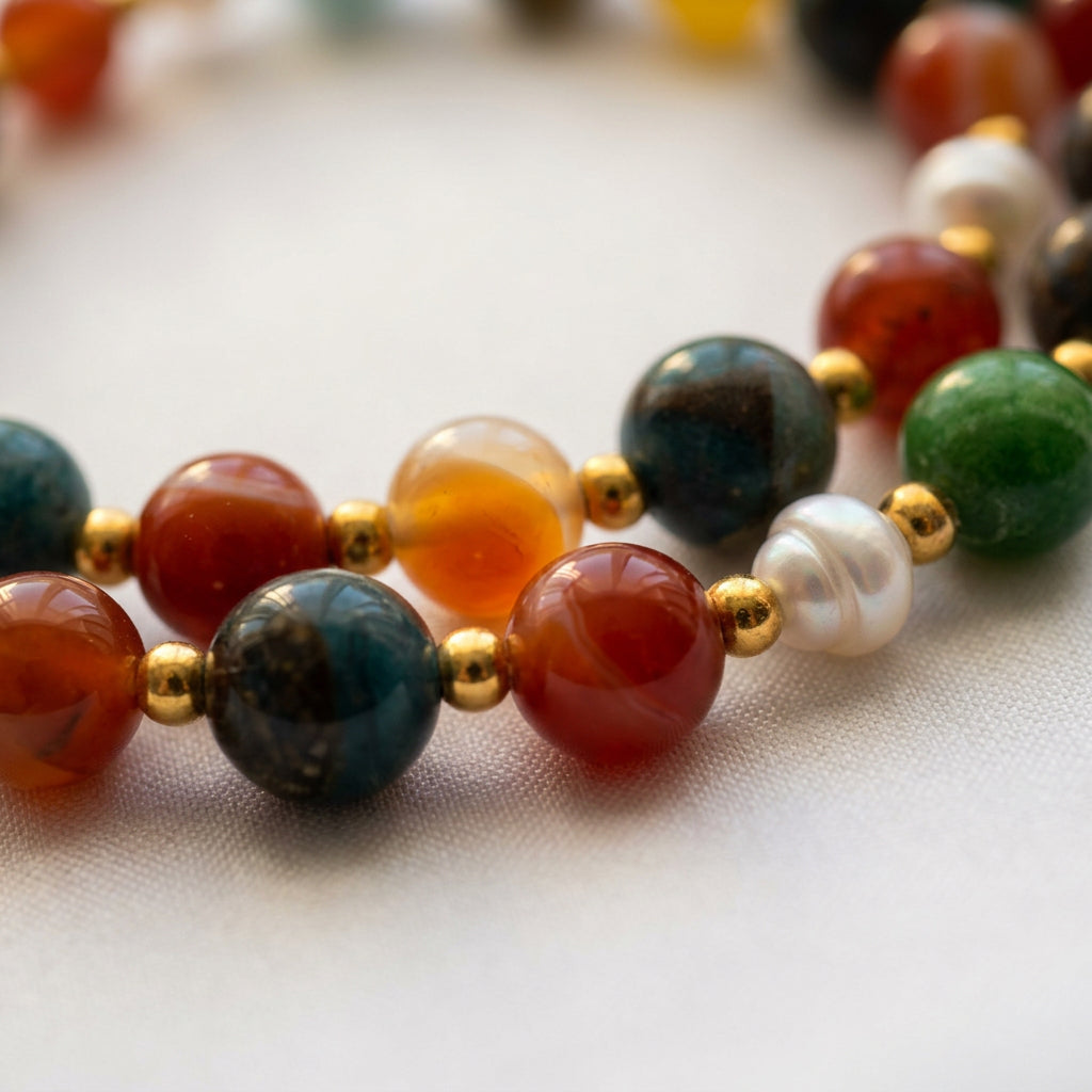 Close-up of blue, orange-red and green stone beads beside a ridged white baroque pearl, separated by gold-coloured spacers on a necklace resting on pale fabric.