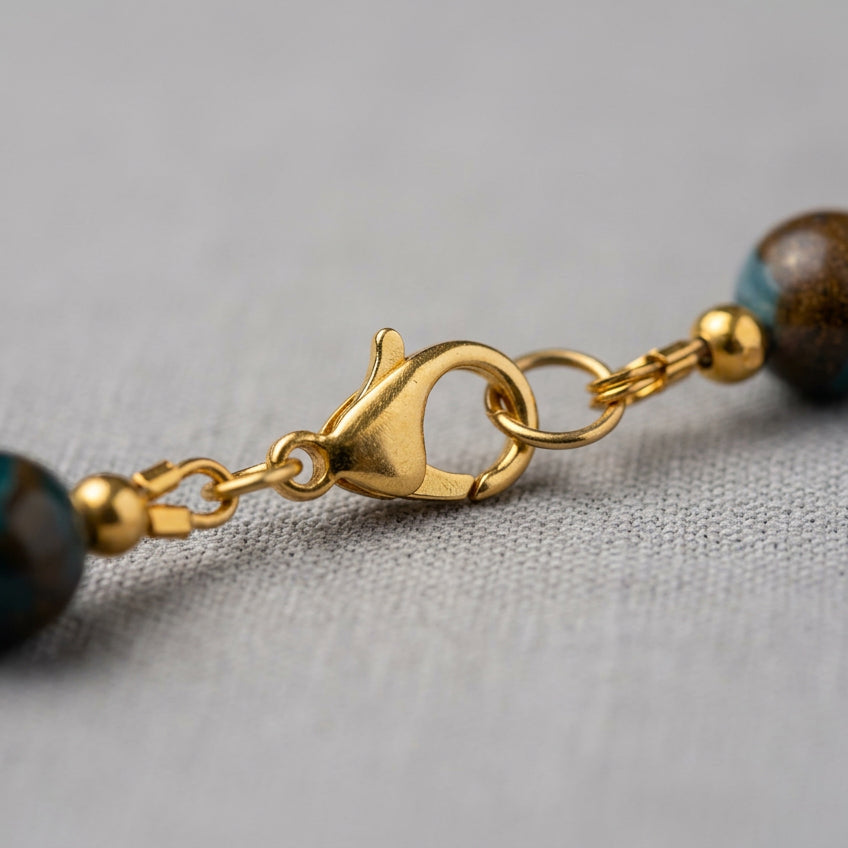 Close-up of the gold-plated stainless steel lobster clasp and connecting rings between blue jasper beads, resting on pale linen.⠋