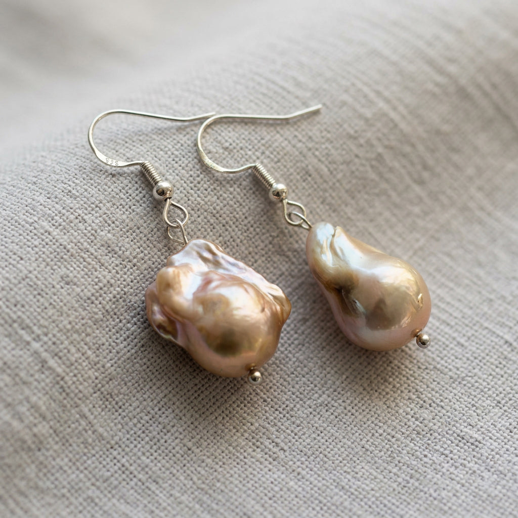 Pair of cream keshi pearl earrings laid on grey linen, showing irregular golden-peach pearls with pink-green iridescence, sterling silver hooks and small silver beads beneath each pearl.
