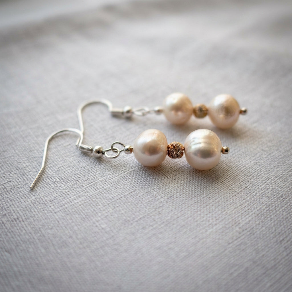 Two pearl drop earrings lying on pale grey fabric, showing the pearls’ natural ridges, textured gold-coloured spacers and curved sterling silver hooks.