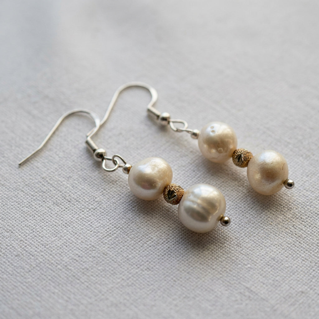 Close-up of double baroque pearl earrings arranged diagonally on grey linen, showing the creamy pearls’ irregular surfaces, patterned gold-coloured spacers and sterling silver hooks.