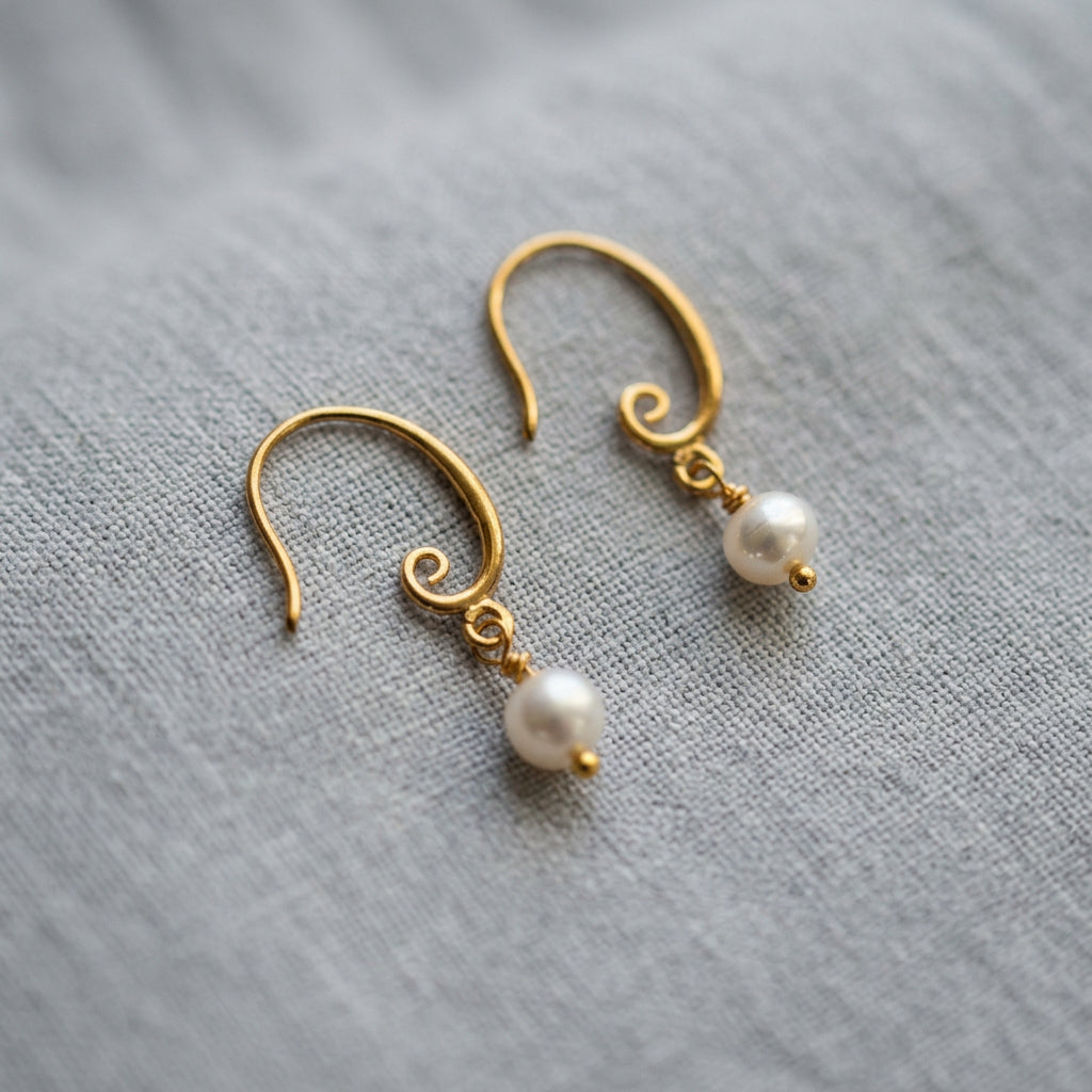 Pair of white baroque pearl drop earrings on pale linen, with curved gold-filled hooks featuring spiral details and small gold bead tips beneath the pearls.