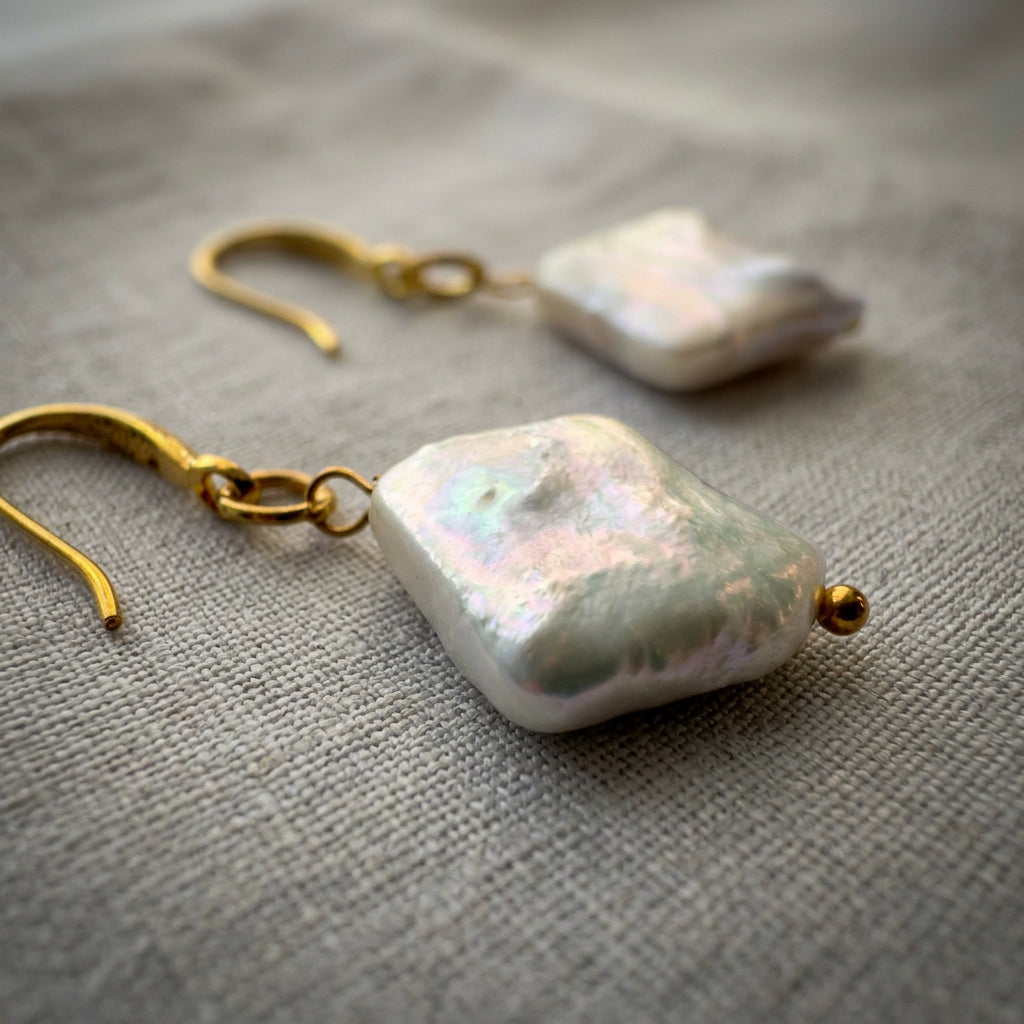 Close-up of white square baroque pearl drop earrings on grey linen, showing textured pink-green iridescence, gold-filled hooks and small gold bead tips.