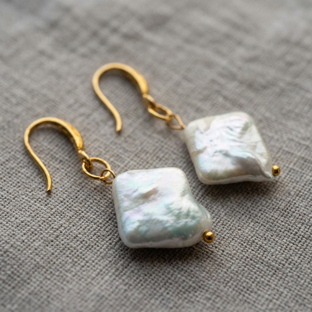 White square baroque pearl drop earrings arranged on cream linen, showing softly rippled nacre surfaces, gold-filled hooks and small gold bead tips.