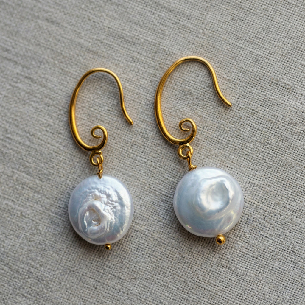Pair of white coin pearl drop earrings on grey linen, showing circular nacre ridges, natural indentations and gold-filled hooks with spiral details.