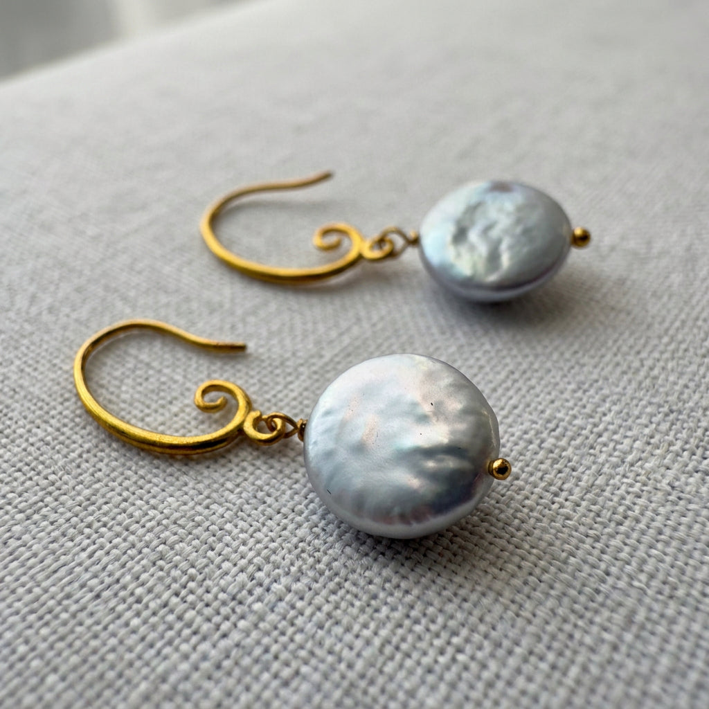 White coin pearl drop earrings on pale linen, showing softly rippled, silvery nacre surfaces, gold-filled spiral hooks and small gold bead tips.
