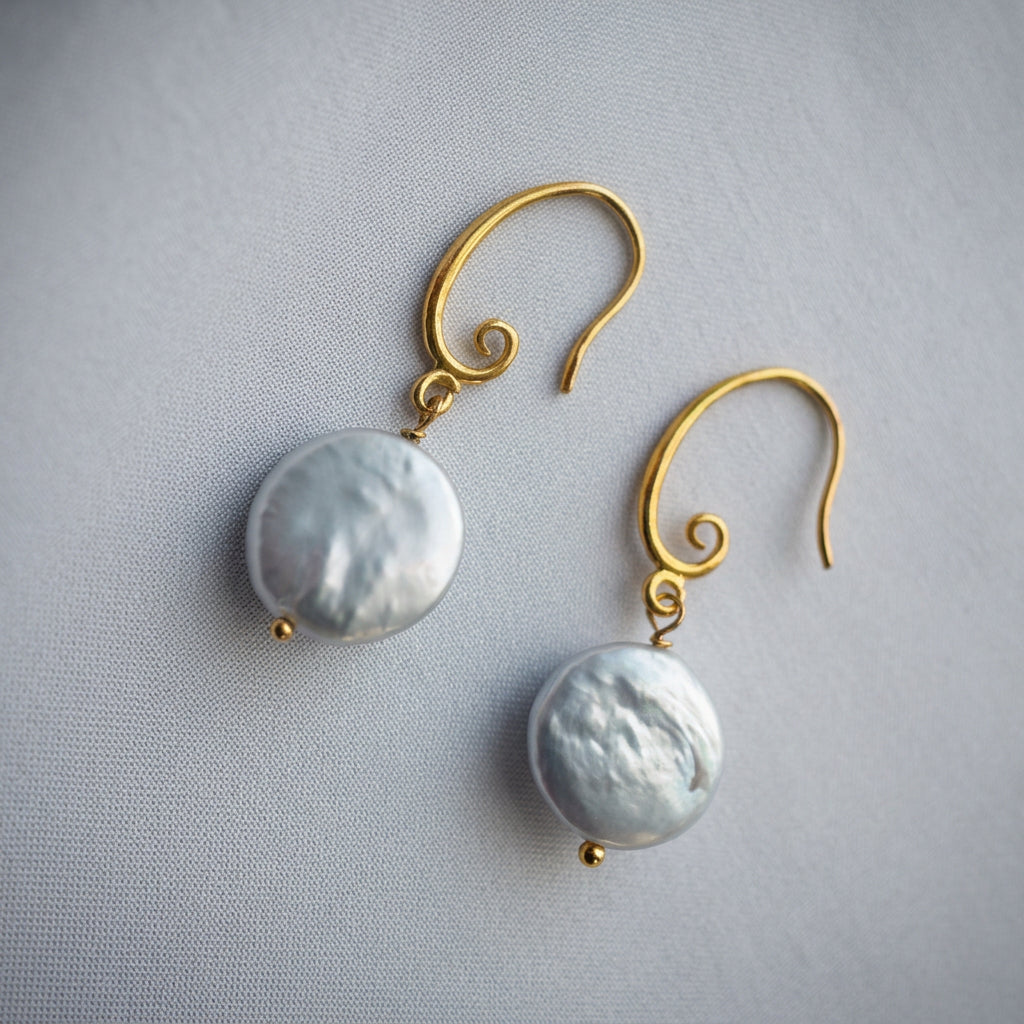 Pair of white coin pearl drop earrings arranged diagonally on pale fabric, showing rippled silvery nacre, gold-filled spiral hooks and small gold bead tips.
