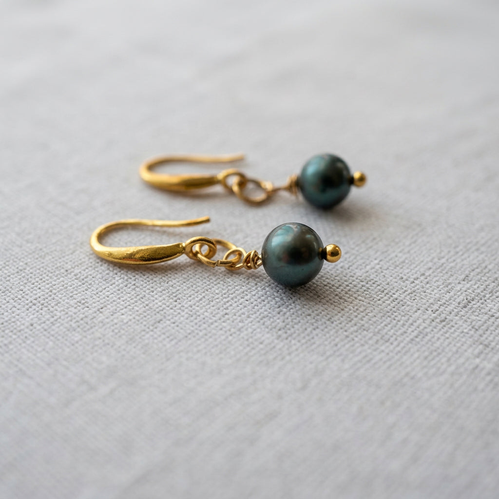 Peacock baroque pearl drop earrings lying side by side on pale linen, with dark green iridescent pearls, gold-filled hooks and small gold bead tips.