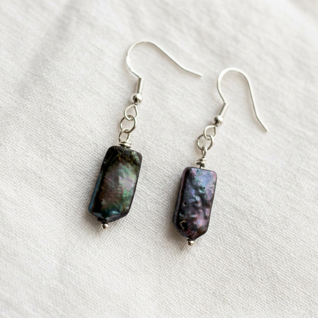 Pair of dark blue square baroque pearl drop earrings on cream fabric, showing green and purple iridescence, sterling silver hooks and connecting rings.