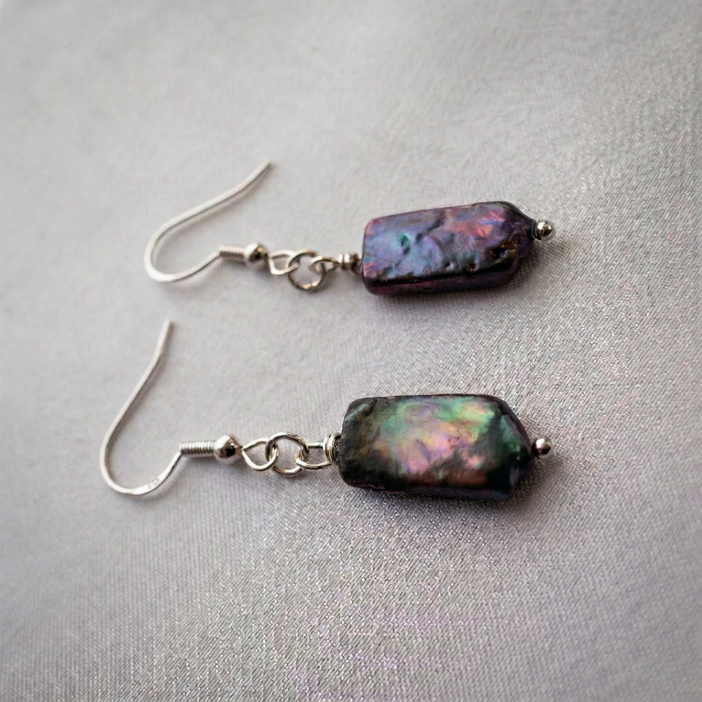 Dark blue square baroque pearl drop earrings on pale fabric, showing vivid green, pink and purple iridescence with sterling silver hooks and connecting rings.
