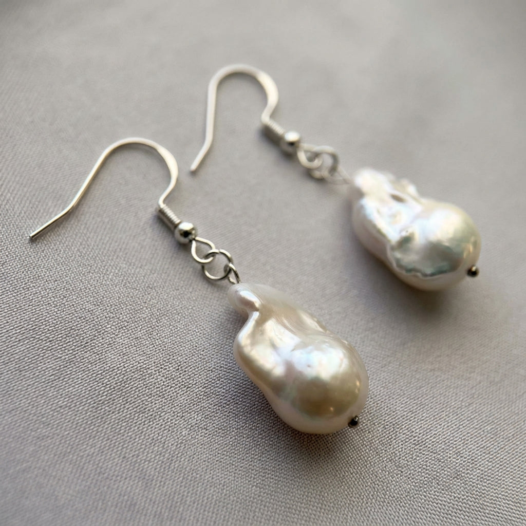 Pair of white Keshi pearl drop earrings on pale fabric, showing irregular elongated shapes, rippled iridescent surfaces and sterling silver hooks with connecting rings.