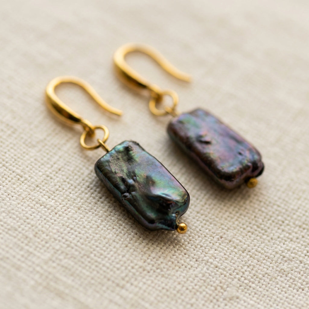 Pair of dark blue square baroque pearl drop earrings on cream linen, showing textured surfaces with green and purple iridescence, gold-filled hooks and small gold bead tips.